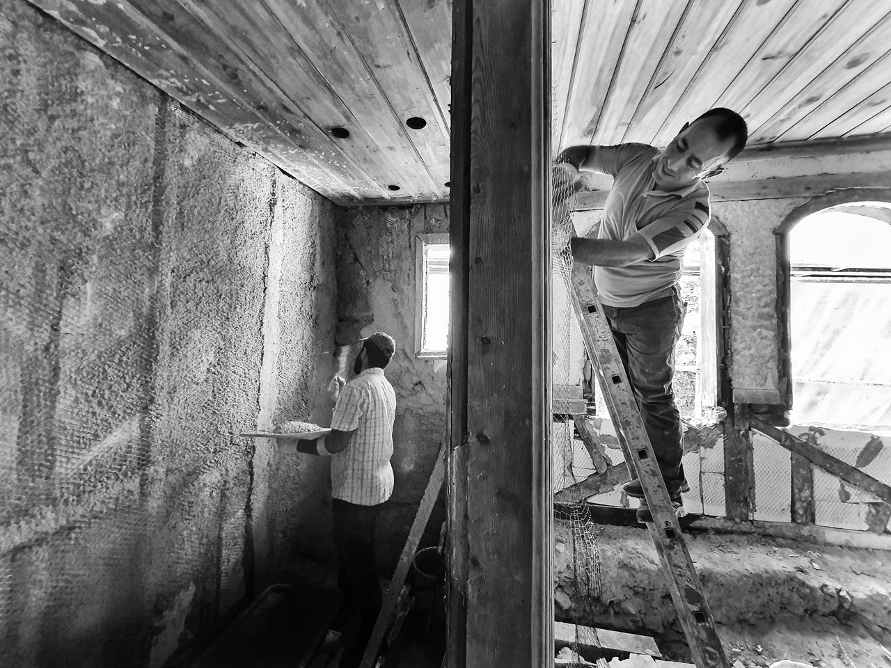about-img-01 Two men renovating a room, one on a ladder applying plaster, showcasing teamwork and effort.